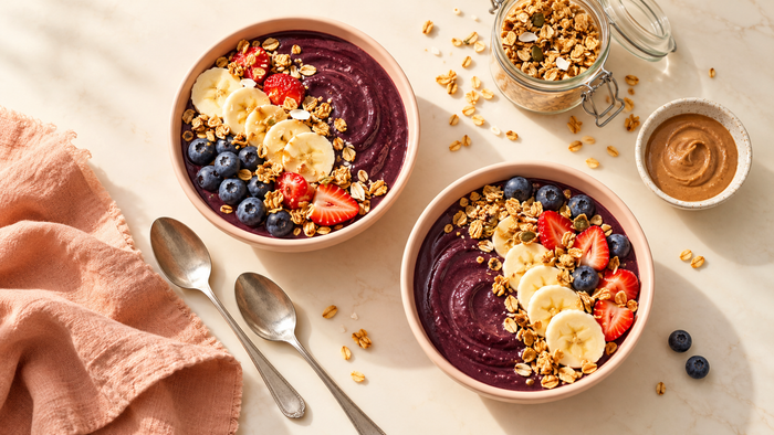 Two açaí bowls on a breakfast table with fruit, granola and nut butter, shown as a serving illustration